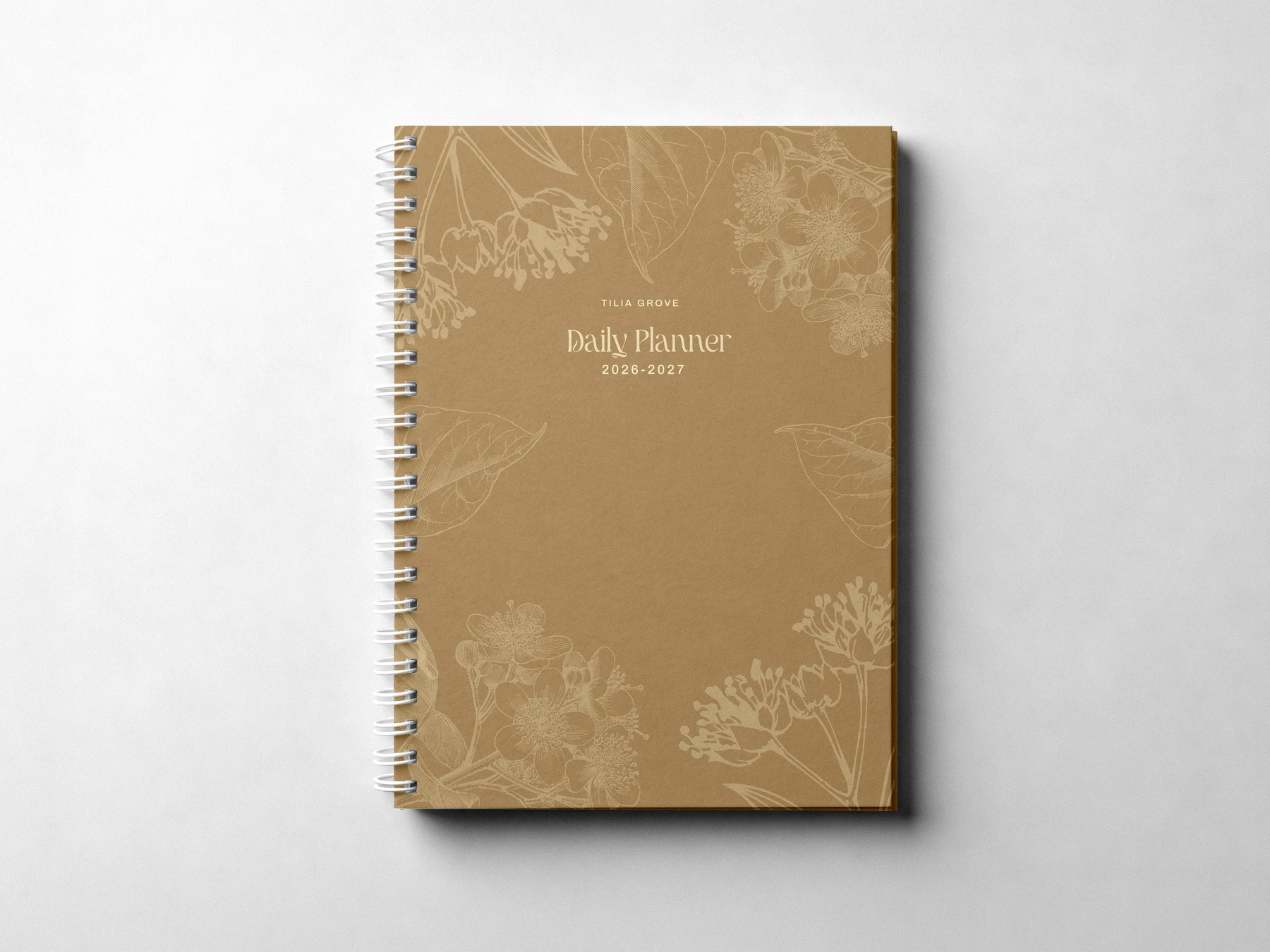 A spiral-bound Tilia Grove 2026–2027 daily planner with a kraft-brown cover and │ │ │ │ planner │ A spiral-bound Tilia Grove 2026–2027 daily planner with a kraft-brown cover and gold botanical illustrations.